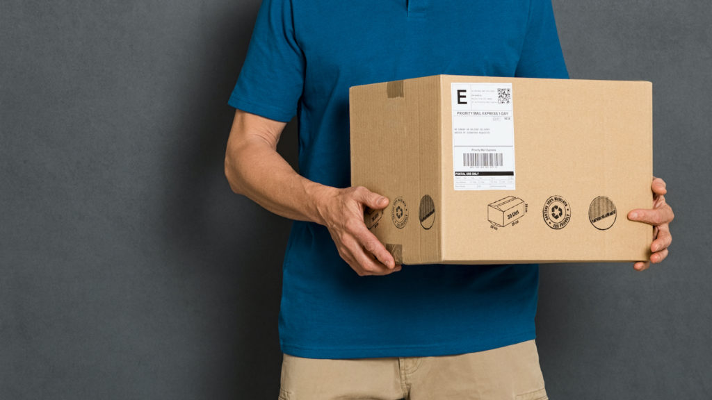 Happy courier holding cardboard box and looking at camera. Smiling delivery man holding and carrying a card box isolated on grey background. Portrait of deliveryman holding parcel with copy space isolated.